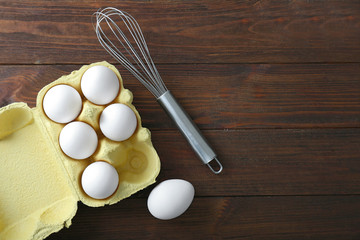 Raw eggs and whisk on wooden background