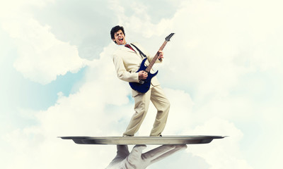 Businessman on metal tray playing electric guitar against blue sky background