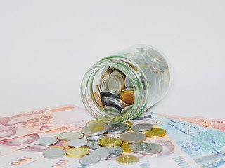 Coins spilling out of glass jar on banknotes and white background