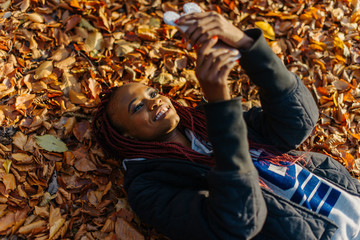 Beautiful african american girl lying in yellow leaves in autumn park. Female making selfi and...