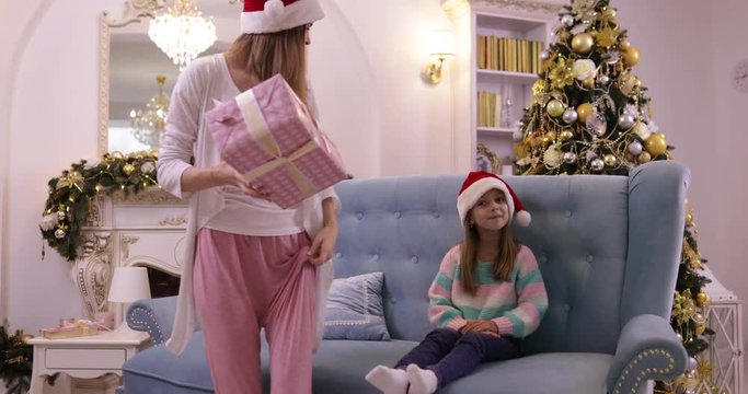 Mother Give Daughter Present Box Gift Happy Smiling Family On Couch Wear Santa Hat Decorated New Year Christmas Tree Small Girl And Woman