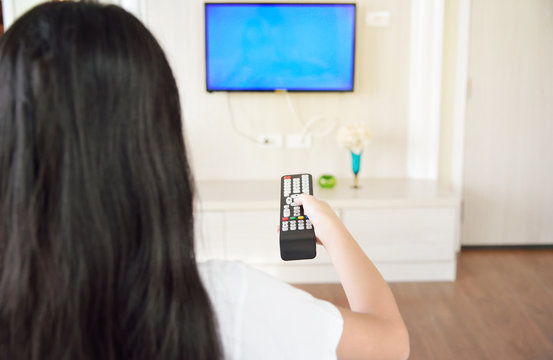 Over the shoulder view of young Asian girl with long black hair sitting on sofa holding tv remote and surfing programs on television. Teenage woman watching TV in the living room