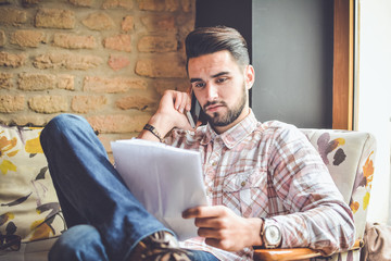 Businessman sitting at table in cafe, reading newspaper and talking on mobile phone