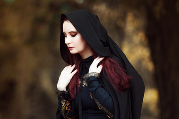 Young beautiful and mysterious woman in woods, in black cloak with hood, image of forest elf or...