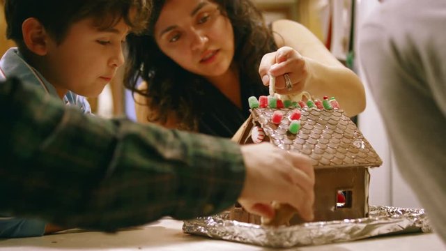 Smiling mom and son decorate a chocolate house at kitchen table with family