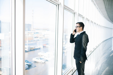 Handsome businessman standing next to the large windows of his top floor office, looking at the view of the city while talking on his mobile phone