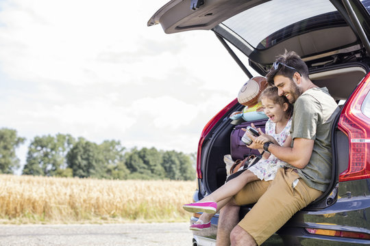 Father and daughter sitting in open car boot looking at camera at break of a road trip