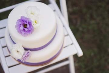 White wedding cake with purple flowers decoration outdoors on wooden modern chair. Top view, copy space.