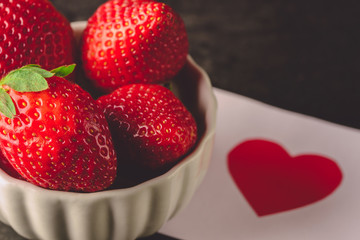 Red fresh strawberry in white bowl on black background and valentines day card with red heart. Valentines day romantic dinner