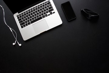 Laptop, smart phone, sunglasses and earbuds on dark, black desk