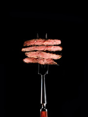 Slices of Medium rare grilled Steak Ribeye on meat fork on black background