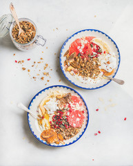 Healthy breakfast yogurt bowls with granola, grapefruit, banana, pomegranate, dried fruit, nuts, seeds over grey marble background, top view. Clean eating, detox, gluten-free, allergy-friendly concept
