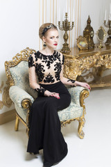 Beautiful blonde royal woman sitting on a retro chair in gorgeous luxury dress, looking at camera. Indoor
