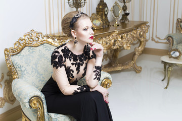 Beautiful blonde royal woman sitting on a retro chair in gorgeous luxury dress. Indoor. Copy Space