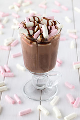 Glass of hot chocolate with colorful mini marshmallows and topping on white wooden table.