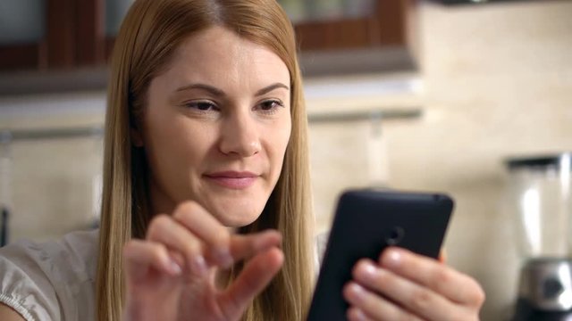 Beautiful young woman with a phablet sitting in kitchen. Browsing Internet and smiling.