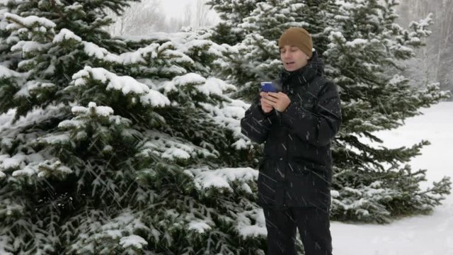 Young man typing a sms message on the phone in the winter snowy forest. Big snowfall. He smiles and speaks the written.