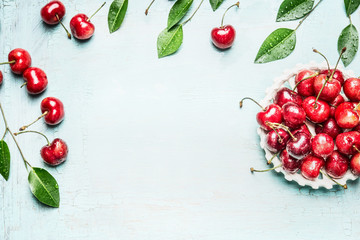 Red ripe cherry  berries in bowl with twigs and leaves on light blue background, top view frame....