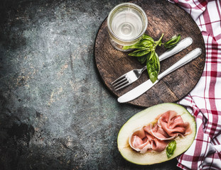 Mediterranean meal: melon with ham , wine and cutlery served on wooden cutting board, top view