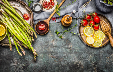 Tasty asparagus cooking with tomatoes, lemon and seasoning, preparation on rustic kitchen table with wooden spoon, top view, border. Dark style - obrazy, fototapety, plakaty