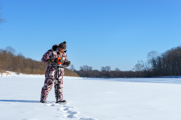 Bearded man is drilling ice hole by automatic moto boer for winter fishing at sunny day under blue...
