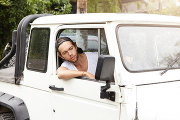 People, lifestyle, travel and tourism concept. Handsome young male tourist wearing snapback driving his white vehicle, enjoying wild nature during safari adventure trip, looking out car window