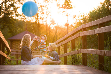 Mother with kid boy and cute little toddler sitting on wooden bridge and playing with blue balloon on summer sunset. Woman with children outdoors. Lifestyle concept
