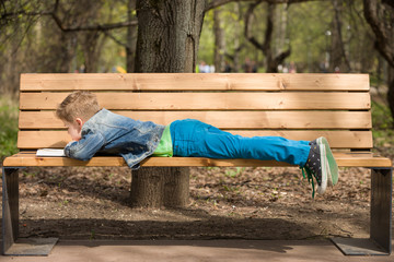 Cute kid boy lying on the bench in the park and reading a book on a sunny summer day. Child learning and reading. Education. Outdoors.
