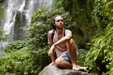 Stylish young Caucasian tourist with backpack relaxing barefooted on big stone with gorgeous waterfall behind him. Bearded backpacker sitting on rock during trip in mountains and looking tired