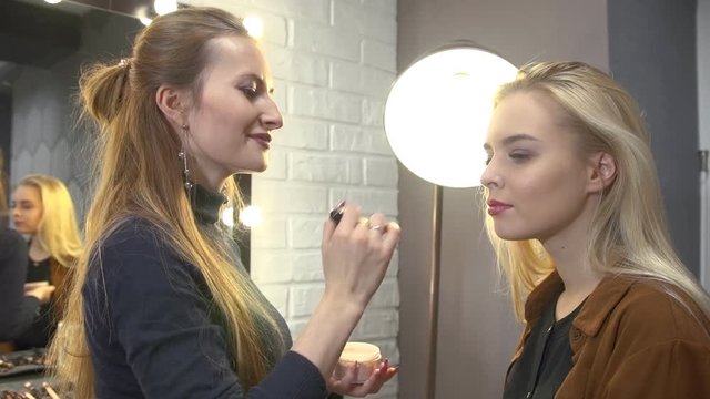 Young  visagiste putting makeup the face of beautiful blonde girl sitting on a chair next to the mirror in the beauty salon.