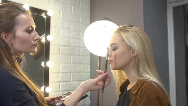 Young  visagiste putting makeup the face of beautiful blonde girl sitting on a chair next to the mirror in the beauty salon.