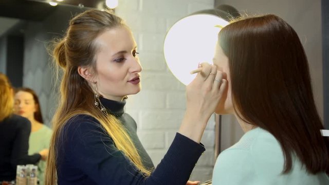 Young visagiste putting makeup on face of beautiful brunette girl sitting on a chair next to the mirror in the beauty salon.