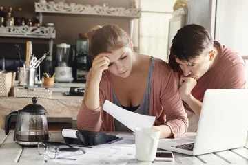 Fotobehang Young married couple facing financial problem during economic crisis. Frustrated woman and unhappy man studying utility bill in kitchen, shocked with amount to be paid for gas and electricity © Wayhome Studio