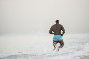 Dark muscular man running down the beach splashing water showing back