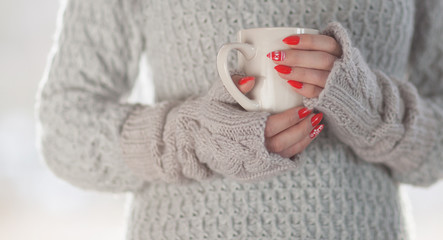 Woman wearing knitted sweater and gloves holding a cup of hot coffee, humorous home heating energy saving