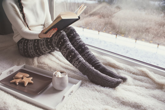 Young woman with cup of coffee and gingerbread is reading a book sitting home relaxing by the window. Snow winter home chill out concept.