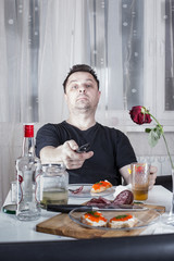 Drunk unshaven man sitting at a table with drinks and snacks, holding a remote control and looks at the camera.