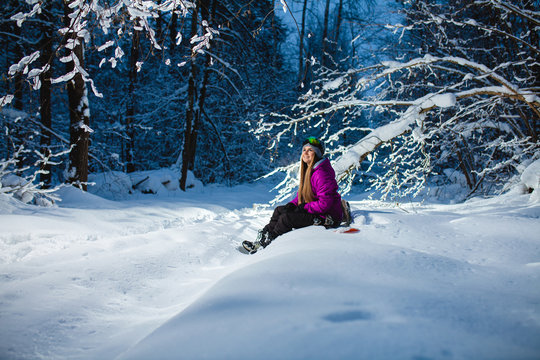 Young sexy woman sitting on her snowboard in the dark winter forest