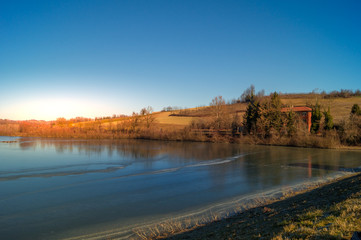 expanse of green grass on the lake with blue sky, countryside and farm around the lake, sunlit water