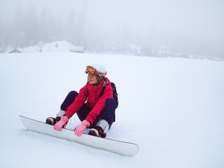 Attractive female snowboarder sitting in snow in winter costume on  mountain. Winter landscape in background.