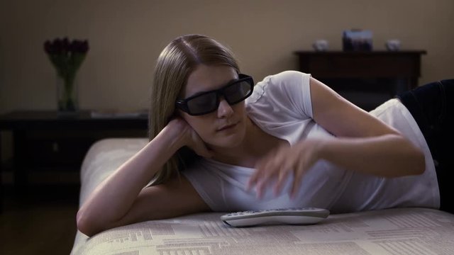 Young beautiful woman lying on couch. Puts on 3d-glasses switches on smart TV and watches 3d movie