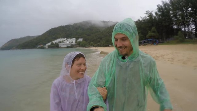 Loving Couple Walking and Smiling on the beach in raincoats. Rain season Thailand. Selfie Action on camera.