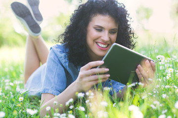 Pretty curly hair woman laying on grass with white flowers around her. She is using a tablet.