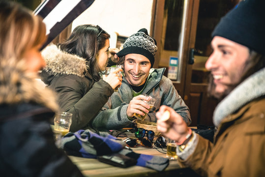Happy friends drinking beer and eating chips after ski - Friendship concept with cheerful people having fun at bar restaurant resort with snow equipment - High iso image with shallow depth of field