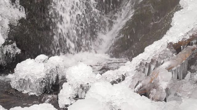 Iciciles bellow waterfall. Snowy and icy stones and boulders with drops of fallen chilly water. Fallen icicle bellow waterfall, stony and snowy stream bank. Close focus
