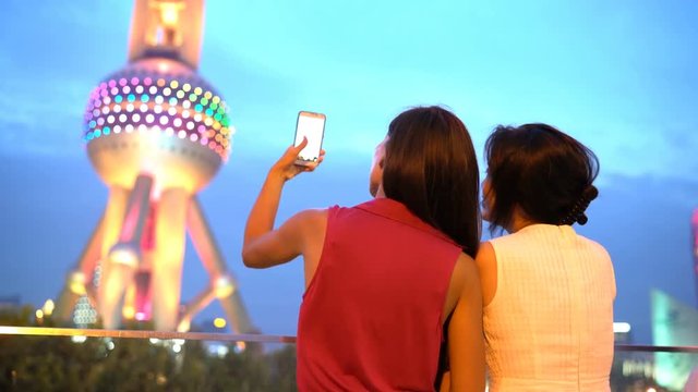 Shanghai China tourists taking photo with smart phone of Oriental Pearl Tower at night at famous chinese tourist attraction destination. Woman friends on travel taking having fun in China.
