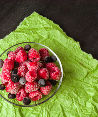 Frozen raspberries in a glass saucer. Frost on the berries. Dark and green background. Green crumpled paper.