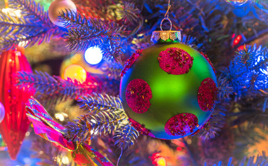 Fototapeta na wymiar Christmas tree decorations. Green, matte finish, orb with magenta circles, glows, surrounded by vivid blue and multicolored mini-lights close up on a small faux indoor Christmas tree. 