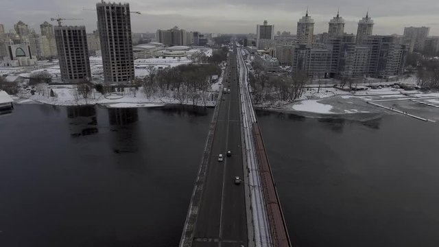 cars go over the bridge, next to moving subway train, on the street it is snowing, nedostoroennoe building stands in the background.