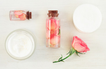 Set of aromatic botanical cosmetic products on white wooden table. Glass bottles with extract, tonic infused with flower petals, fresh pink rose, cotton pads. Soft light and focus.